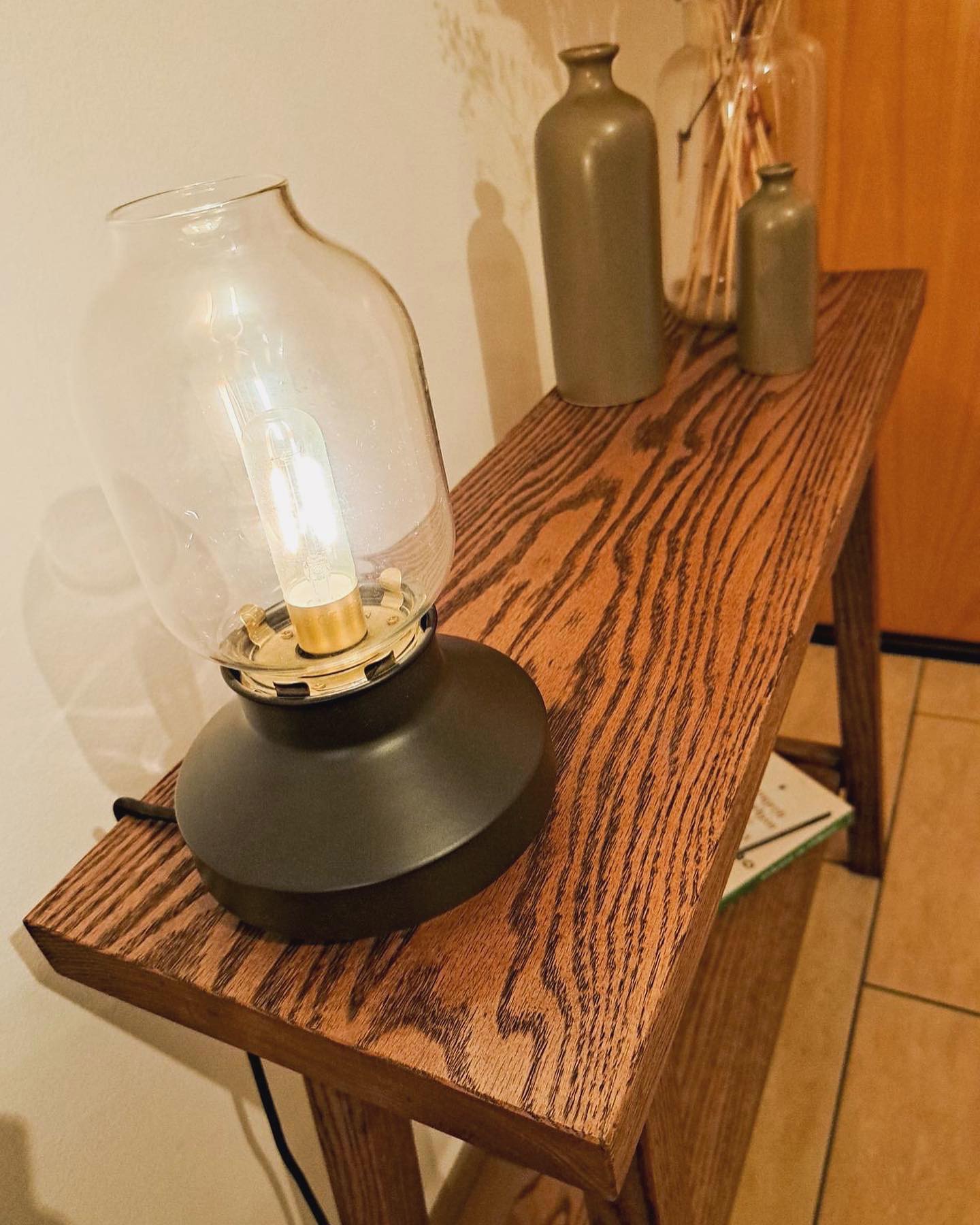 The lamp lit on the table top, its glass shade glowing above the open grain of the oak