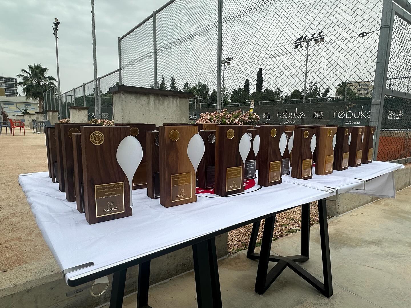 The full set of trophies laid out on a trestle table beside the tennis courts under a grey sky