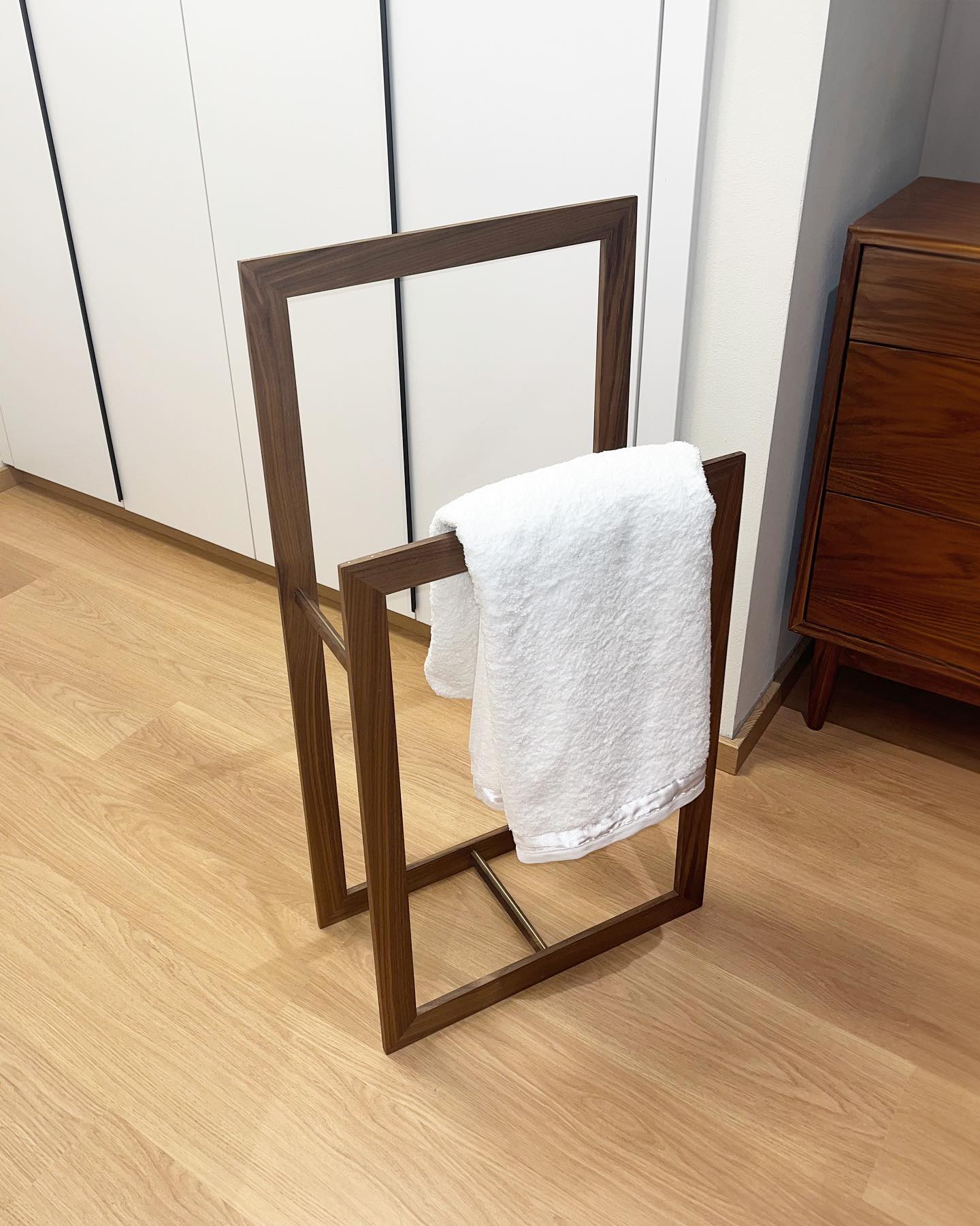 The same valet on a pale wood floor beside a chest of drawers, a white towel hanging from the front frame